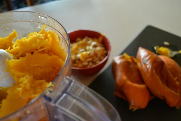 Pumpkin pulp in a food processor, with pumpkin seeds in a bowl and empty cooked pumpkin shells in the background.