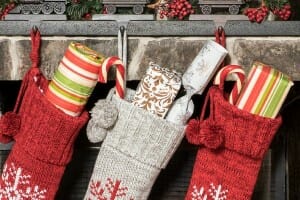 Three sweater knit stockings with tassels hung on a fireplace mantel, filled with wrapped presents.