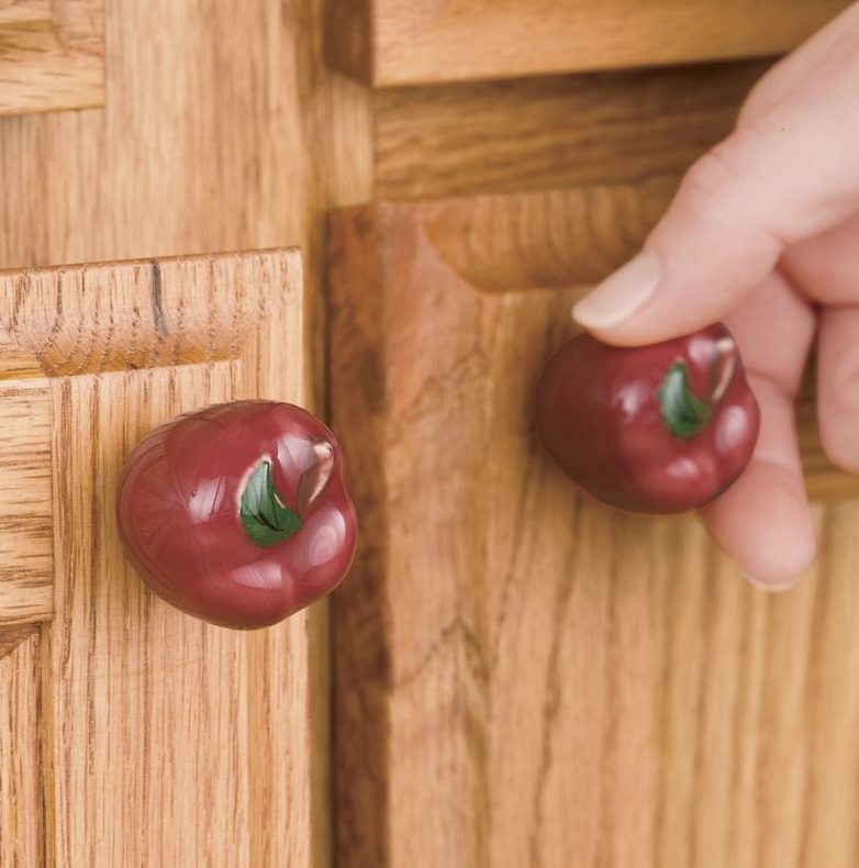 Close-up of a hand opening a wood cupboard door with red apple knobs.