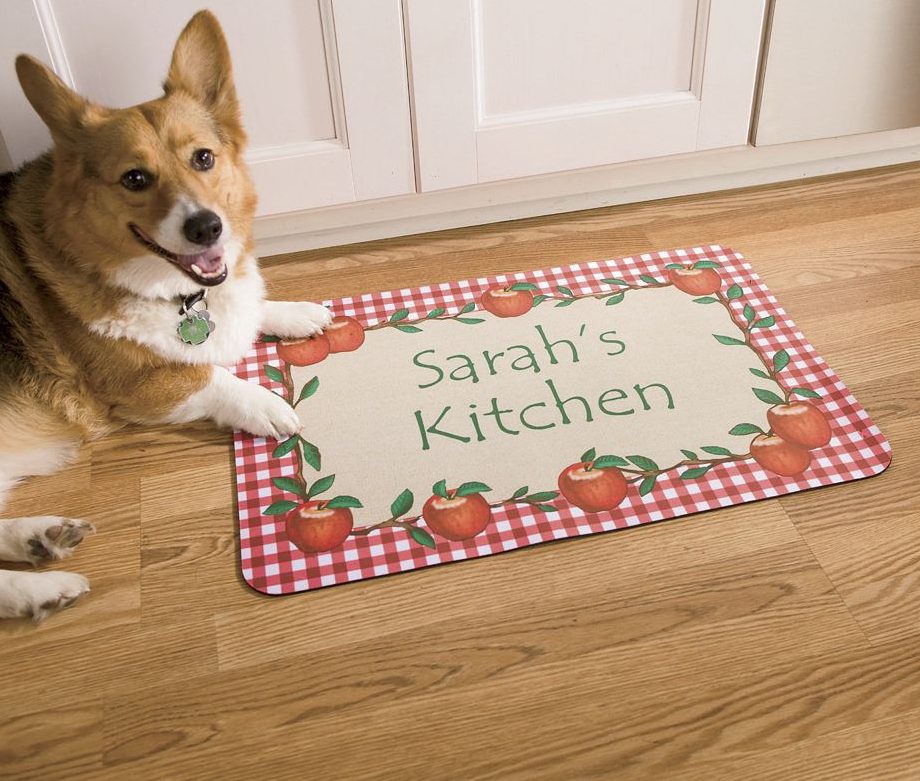 A brown and white Corgi dog lying by a personalized door mat with a border of red and white checks and red apples.