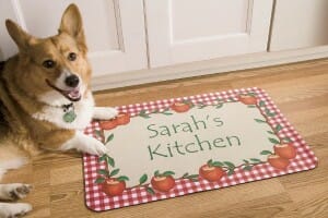 A brown and white Corgi dog lying by a personalized door mat with a border of red and white checks and red apples.