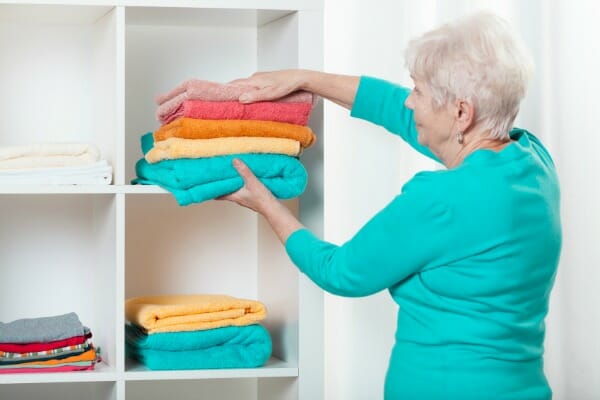 A silver-haired woman in a teal top putting a folded multicolor stack of towels into a storage cube.