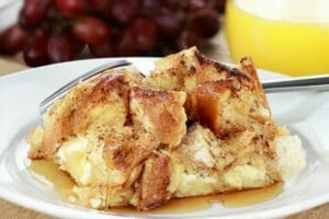 A white plate with a serving of French Toast Casserole, with a fork and fresh red grapes.