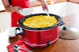 A woman ladeling a serving of Macaroni and Cheese into a white bowl, from a red Hamilton Beach slow cooker.