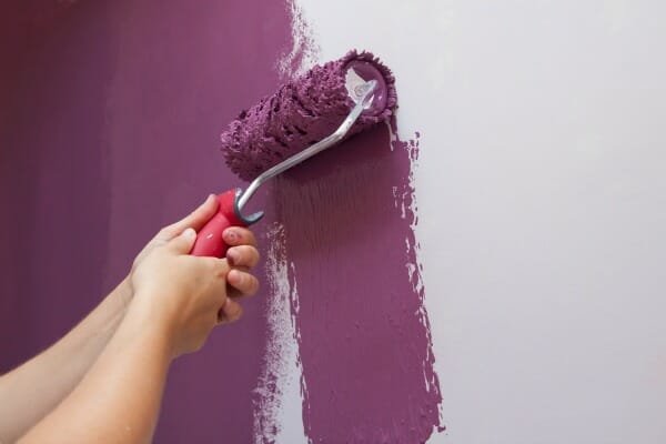 Close-up of a person using a roller to paint a white wall a plum color.