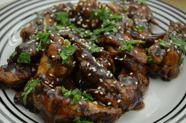 A white plate with black stripes, filled with Asian chicken wings topped with sesame seeds and shredded parsley.