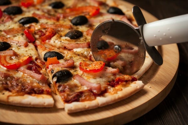 A Supreme pizza on a round cutting board, with a pizza cutter cutting a slice.
