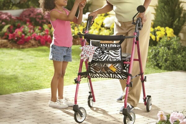 A young girl with an older woman holding onto a pink and black walker with a zebra print supplies holder.