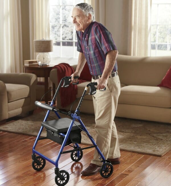 An older man in a living room, holding on to a blue and black walker with hand brakes.