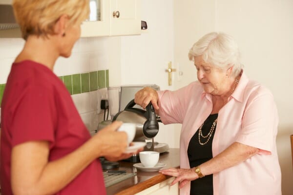 A blonde woman in a kitchen with a silver-haired woman pouring a cup of coffee.