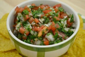 A green and white bowl filled with chunky Pico De Gallo, placed on a plate filled with corn chips.