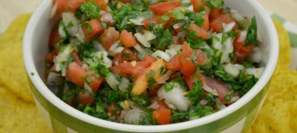 A green and white bowl filled with chunky Pico De Gallo, placed on a plate filled with corn chips.