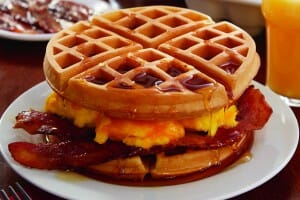 A Waffle Breakfast Sandwich with scrambled eggs and crisp bacon in the middle, topped with maple syrup.