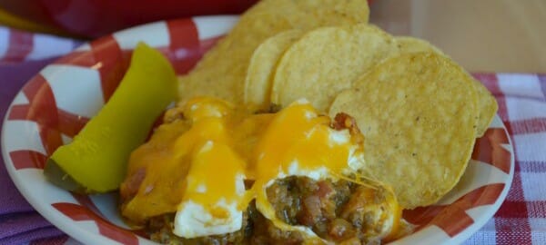 A red and white bowl filled with corn chips, a dill pickle, and Bacon Cheeseburger Dip topped with melted cheese.