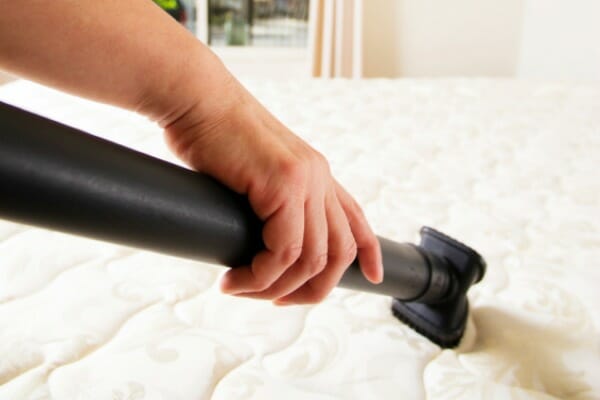 A hand holding a black vacuum hose, cleaning the top of a white mattress.