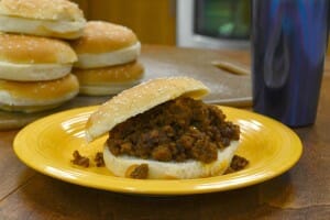 A yellow plate with a Sloppy Joe sandwich, and a stack of buns in the background.