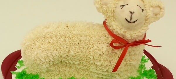 A white lamb-shaped cake with fluffy white frosting, with a red ribbon around the neck and placed on green coconut grass.
