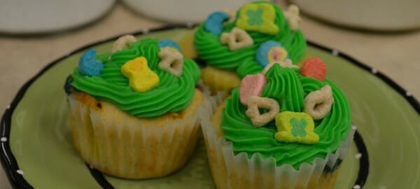 Three cupcakes on a green plate, decorated with green frosting and Lucky Charms cereal and marshmallows.
