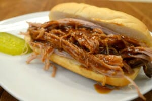 A white plate with barbeque pulled pork on a hard bun, with a dill pickle slice on the side.
