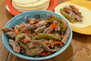A blue bowl filled with Slow Cooker Fajitas next to a stack of soft taco shells, and a plate with a single serving.
