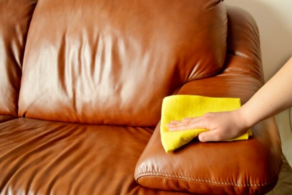 A person's hand using a yellow cloth to clean a brown leather sofa.