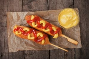Two corn dogs on sticks, covered with swirls of ketchup and mustard, placed on wax paper next to a bowl of mustard.