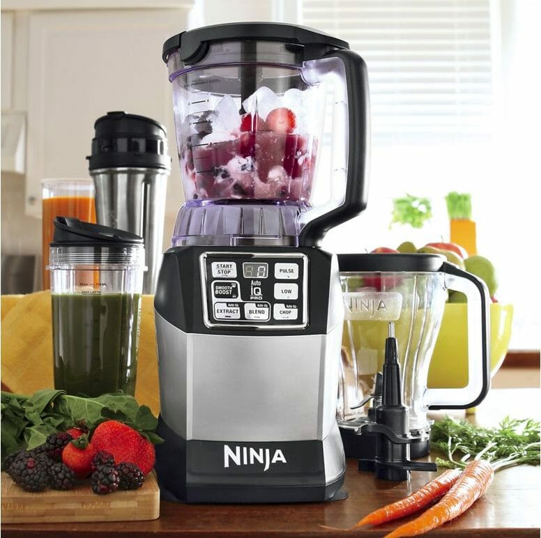 A berry filled Ninja food processor with accessories, including a blending cup filled with a green beverage.