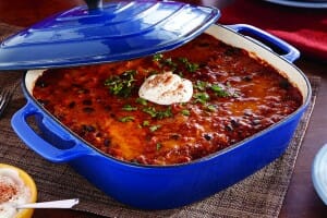 A square blue casserole pan with a matching cover, filled with Enchilada Casserole that's topped with sour cream.