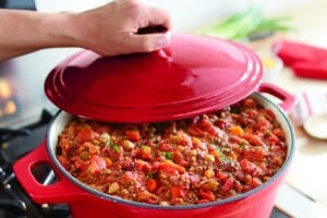 A person lifting the lid of a covered red casserole pan that is filled with Lazy Day Stew.