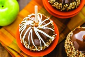 Three caramel apples with nuts in orange dessert bowls, one with white icing swirls, and a Granny Smith apple.