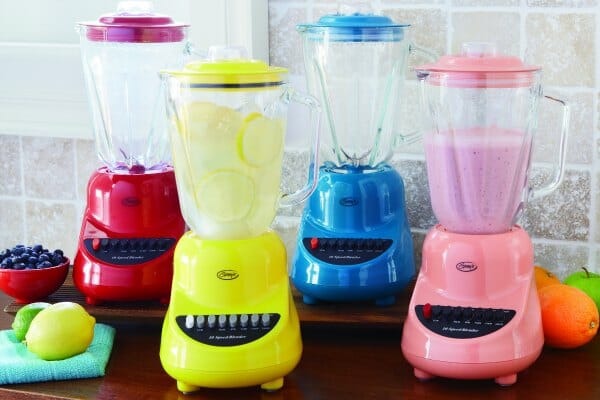 Four Ginny's brand blenders on a counter with fruits, in red, yellow, blue and pink.