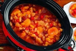 A red and black slow cooker filled with a fruit soup, including apricots, pineapple, and cherries.