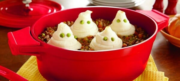 A red pot filled with a casserole and topped with four mashed potato ghosts with green pea eyes.
