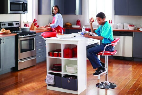 A modern kitchen with a reading black boy on a red chrome stool next to a white island, with his smiling Mom looking on.