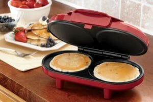 Two blueberry pancakes in a red and black electric pancake maker, with a stack of pancakes on a plate with fruit.
