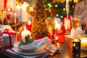 A holiday party table lit with white candles, a Christmas tree centerpiece, white dinnerware and champagne stemware.