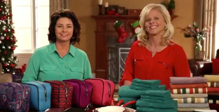 Two smiling women and a table covered with Holiday gifts, including towel sets, pan sets, and cosmetic bags.