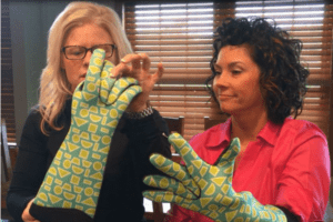Two women putting on long blue patterned oven mitts.
