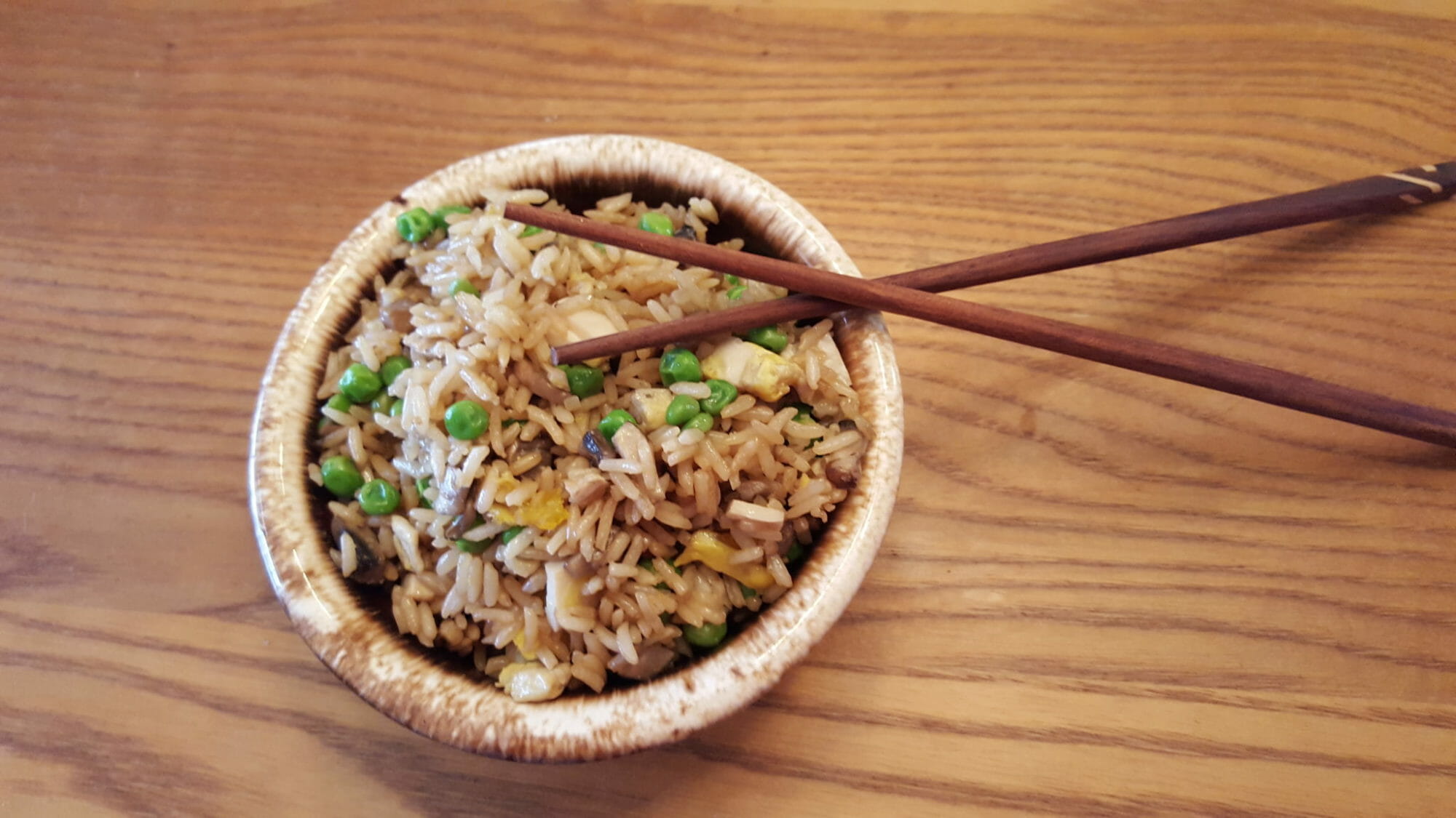 A brown bowl filled with fried rice and vegetables, next to a set of brown chopsticks.