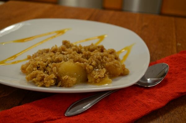 A serving of Apple Crisp with drizzles of caramel on a white plate, with a spoon on a red napkin.