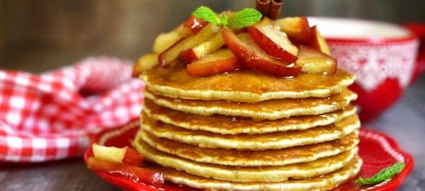 A stack of Apple Pancakes, topped with apple slices, maple syrup, a mint sprig and a cinnamon stick.