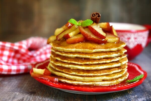 A stack of Apple Pancakes, topped with apple slices, maple syrup, a mint sprig and a cinnamon stick.
