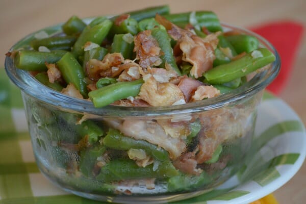 A clear glass ramekin filled with Bacon Green Beans, placed on a green and white plate.