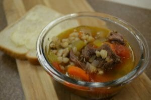 An individual serving of beef barley soup in a clear glass bowl, with a slice of buttered bread.