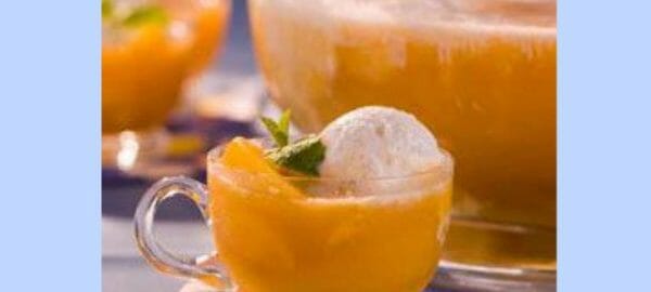 A clear mug of orange Candlelight Punch by a punch bowl, with peach slices, vanilla ice cream, and a mint sprig.