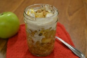 A serving of Apple Crisp in a clear jelly jar, with a spoon and a green Granny Smith apple nearby.