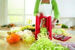 A woman churning out mounds of zucchini noodles using a red spiralizer, with bowls of other veggie spirals nearby.