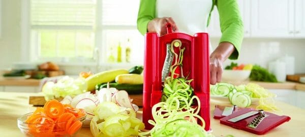 A woman churning out mounds of zucchini noodles using a red spiralizer, with bowls of other veggie spirals nearby.
