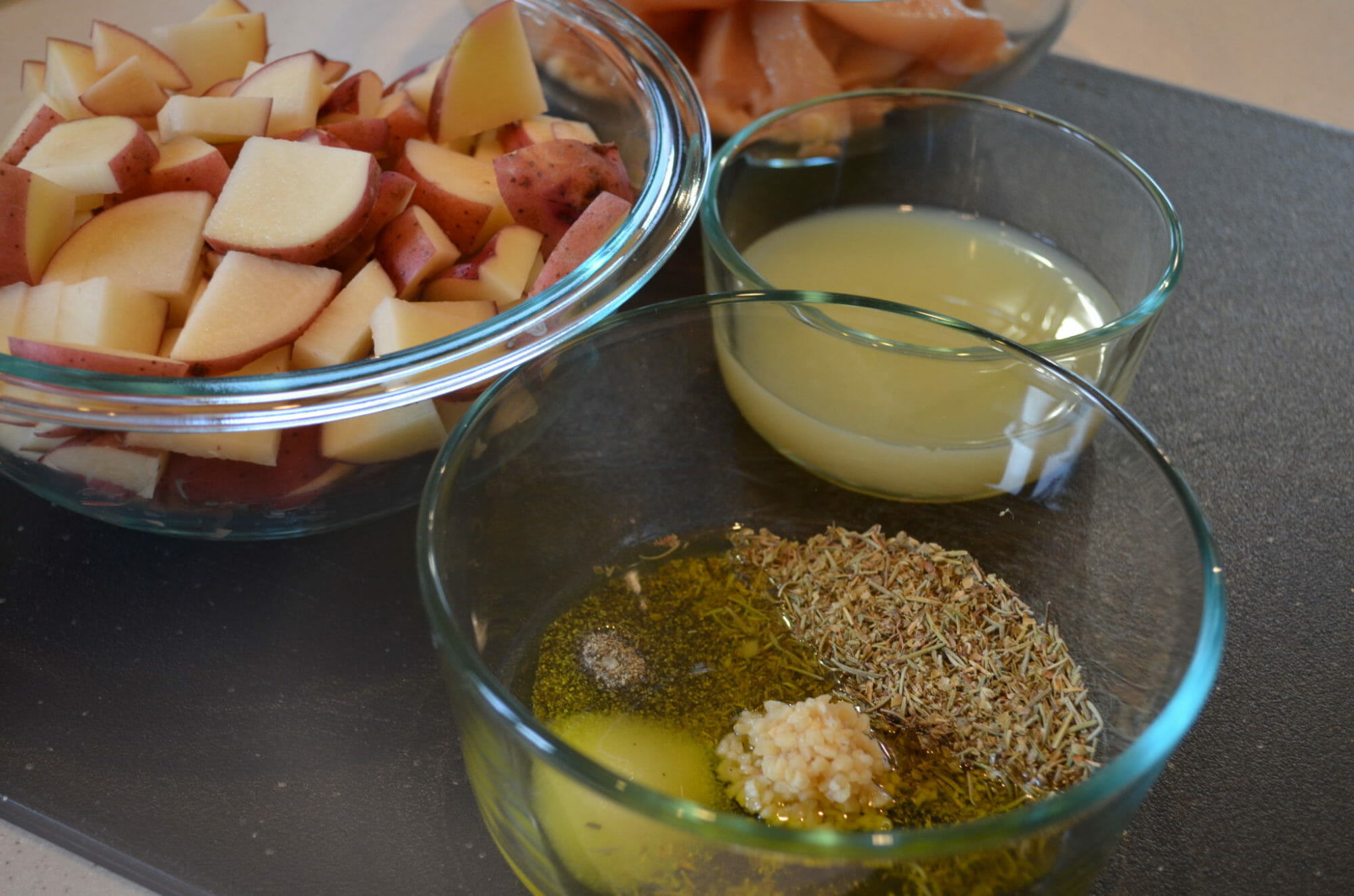Three glass mixing bowls, one with chunks of red potato, one with lemon juice, one with herbal marinade ingredients.