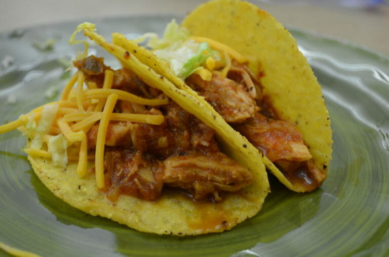 Two slow cooker Chicken Tacos on a green plate, topped with shredded cheese, lettuce, and sour cream.
